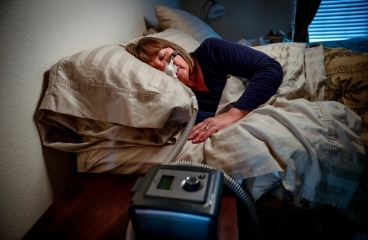 Person wearing nasal pillow that is attached to CPAP machine placed next to bed.